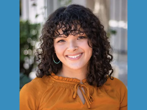 Headshot of woman with brown curly hair and bangs wearing a rust top smiling at the camera.