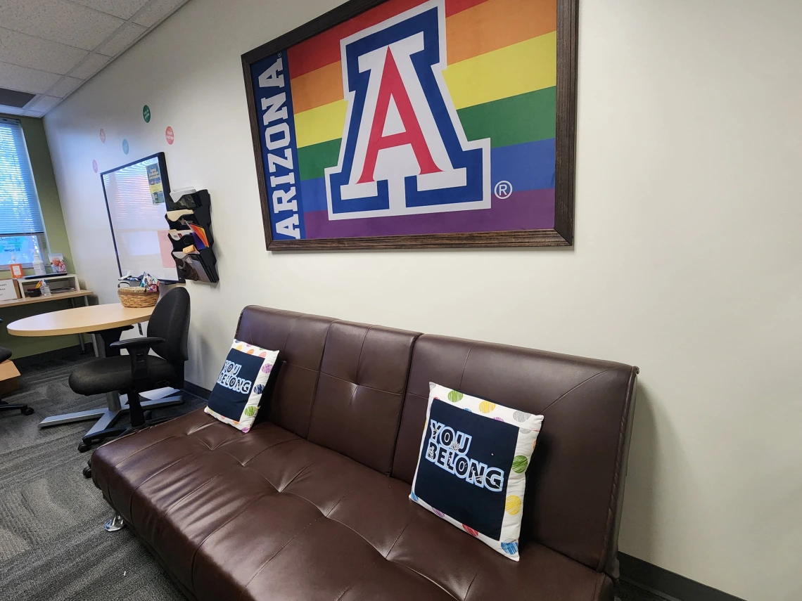 couch with two pillows, small circle desk with two roller chairs, and a UA rainbow flag above the couch