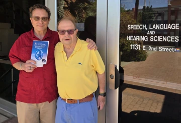 Image of two men standing outside of building doors wearing sunglasses and smiling at the camera.